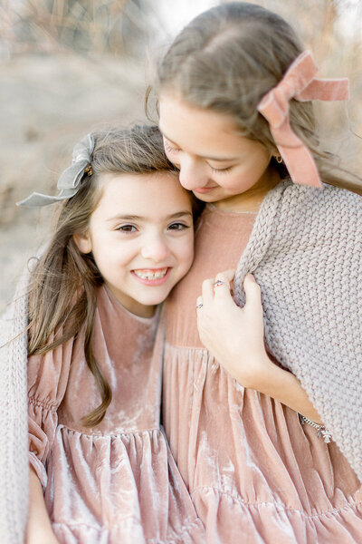 Two young sisters dressed in blush velvet dresses snuggled together in a field under a knitted grey blanket taken in a Dallas/Fort Worth park by a family photographer in Dallas.