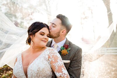 Bride in long sleeve lace dress and groom in tweed suit snuggled together during fall golden hour with veil flowing