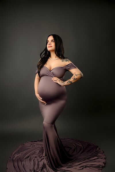 Charlotte area mother to be poses in a tight purple dress with both hands on her belly and posed on a grey waxhaw studio background