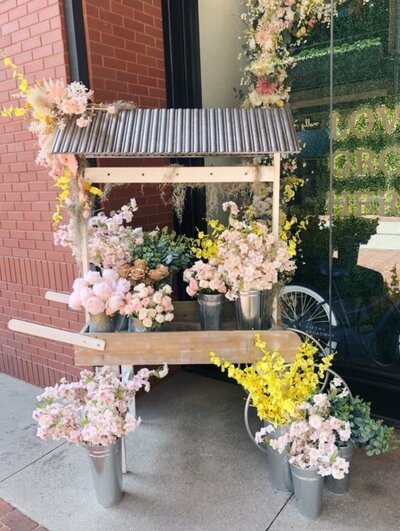 Spring silk floral installation of floral shop cart. Fun spring floral colors such as pink, yellow, white, and green. Retail space floral cart. Design by Rosemary and Finch in Nashville, TN.