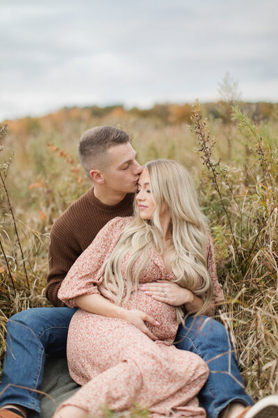 Expecting parents cuddling in field expecting father kissing soon-to-be mother