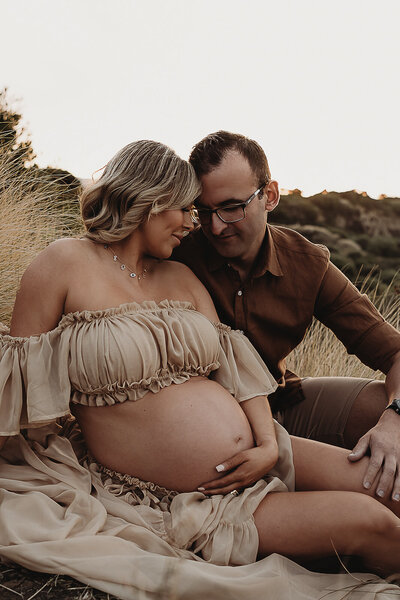 A pregnant woman in a floral dress sits on the ground, cradling her belly in a forested area, captured beautifully by a Wollongong maternity photographer.