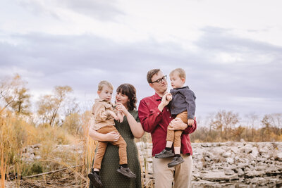 A family of four standing outdoors near rocks and tall grasses with a cloudy sky.