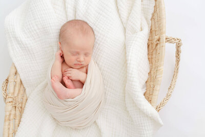 Baby girl laying on a white textured blanket and partially wrapped in a white cozy blanket around her back. Her hands and legs are out from the wrap. Her hands are up by her face and she's got a little pouty face on with a white bow on her head.