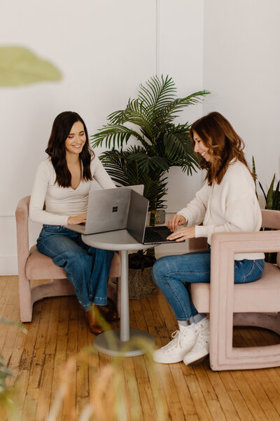 2 women with dark hair in white tops and jeans, sitting in pink chairs with laptops
