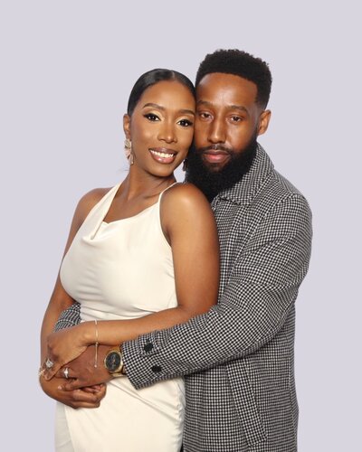 Smiling couple posing at a photo booth, with the woman in a white dress and the man in a houndstooth-patterned coat, embracing in front of a white backdrop.