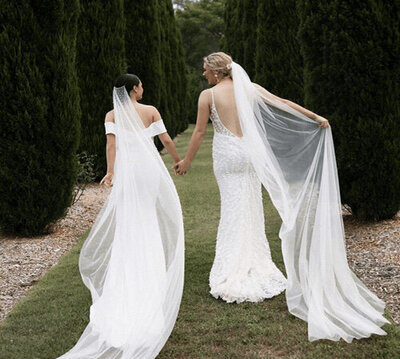 Two brides in stunning white gowns and veils walking hand in hand through a lush garden path