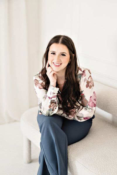 Karli Dawson, assistant and second photographer at Kari Dawson + Co, seated in a relaxed pose wearing a floral blouse and navy trousers, smiling with creative confidence and emotional warmth