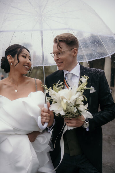 Unter einem Regenschirm geht dieses Hochzeitspaar trotz schlechtem Wetter glücklich nebeneinander her.