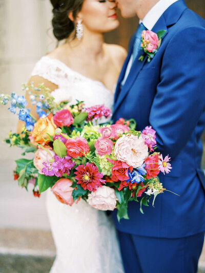 Vibrant rainbow colored wedding bouquet with bride and groom kissing in the background