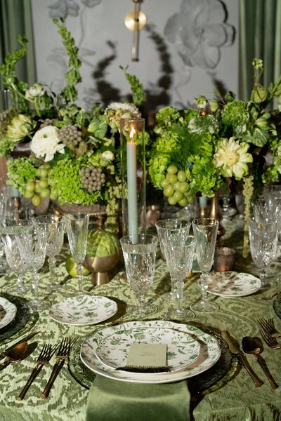 Detail photo of a wedding reception dinner table, with detailed china plates and silverware, green and white floral arrangement, and candlesticks