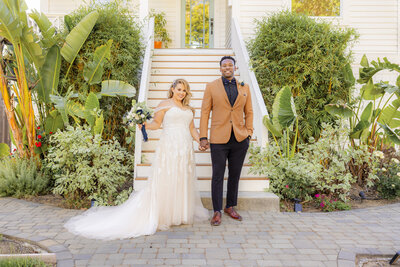Bride and groom stand in front of a porch with white steps looking at camera smiling having just been married.  There is green shrubbery on their sides. Photo taken in midtown Sacramento by wedding studio  photographer, Philippe Studio Pro