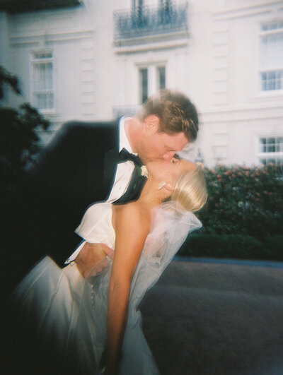 Hazy film photo of bride and groom kissing with white building in the background