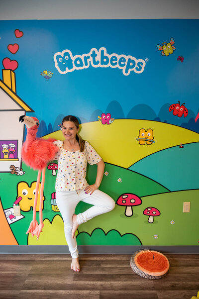 Female, small business owner, standing in front of a Hartbeeps wall mural, posing with one leg on her knee, holding a pink flamingo puppet.