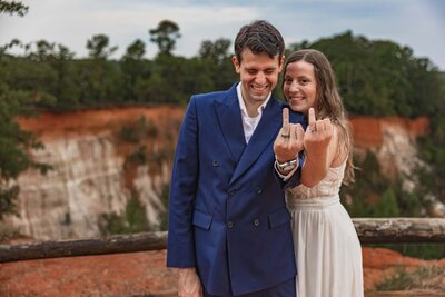 A bride and groom stand in front of the camera holding up their ring fingers to show their wedding rings after just eloping. The bride is standing with her chin resting on the grooms shoulder and smiling at the camera. The groom is laughing, looking down. They are standing in front of a red and orange canyon.