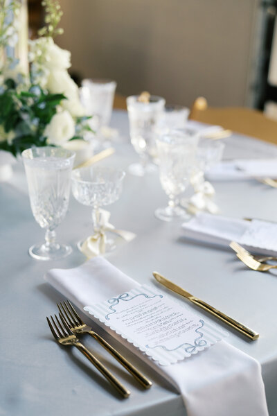 Sophisticated wedding table setting featuring crystal glassware, gold flatware, white floral arrangements, and a personalized menu on a crisp white napkin.