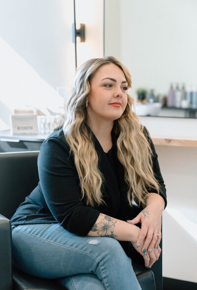 Ingrid, founder of Lavender & Ink Studios in New Franken, WI, seated in salon chair with long blonde hair and tattooed arm