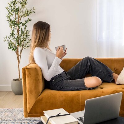 Young woman sitting on a brown velvet couch holding a coffee mug with a laptop on the table, facing toward the subheadline text.