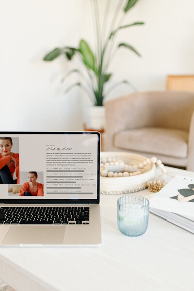 Ein Laptop, der auf einem hellen Holztisch steht. Der Bildschirm zeigt eine Website mit einem professionellen Profil einer Selbstständigen, die ihre Dienstleistungen präsentiert. Auf der Website ist ein großes Profilfoto, eine kurze Beschreibung der Person und ein Menü mit Optionen wie 'Über mich' und 'Leistungen' zu sehen. Neben dem Laptop steht eine dekorative Pflanze und ein Glas.