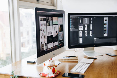 This image displays a professional workspace with two large desktop monitors set up on a wooden desk. The monitor on the left shows a website or a graphic design interface with various thumbnails, while the monitor on the right displays a more text-heavy interface, likely a coding or design application. In front of the left monitor, there's a decorative figurine, possibly a maneki-neko (Japanese lucky cat), accompanied by a small potted plant. The desk also holds a wireless keyboard, a mouse on a mousepad, a notebook, and a smartphone. The environment suggests a creative or tech-oriented workspace, with an urban view visible through the window behind the monitors.