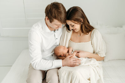 A newborn is laying in his mothers hands while both mum and dad are smiling and looking down on the baby. Captured by Lisa Lefevre Photography