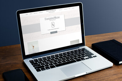 The Fontainebleau Showit Template by Lily is on the screen of a silver laptop sitting on a wood desk in front of a blue wall.