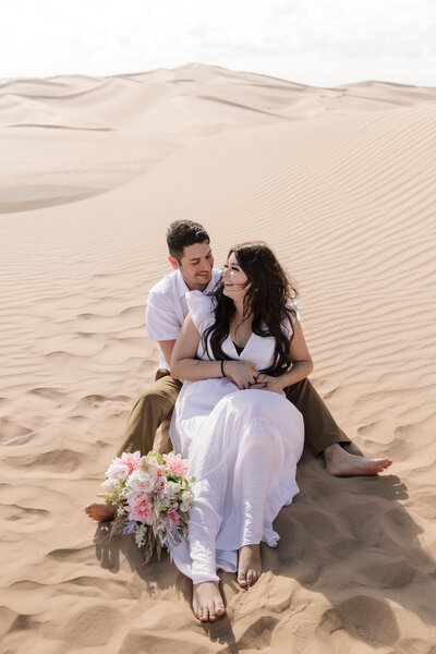 Boho couple wearing neutral color outfits holding each other looking off into the distance at the Sand Dunes