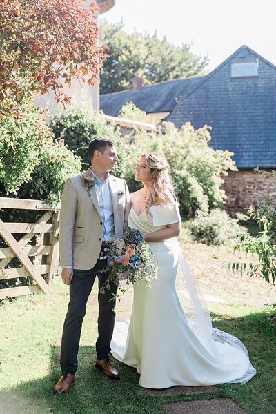 Bride and groom standing in a lush garden with a rustic house in the background, smiling at each other during a countryside wedding