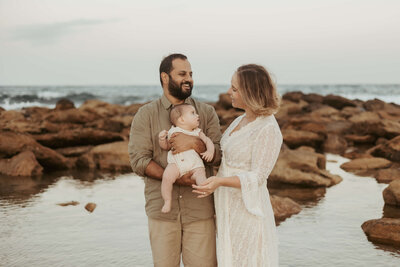 Mum and dad at the beach celebrating their six months of being parents with their baby boy while the ocean and rocks are behind them.