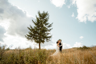 a couple posing in a fild, with green pines trees in the background. The bride is wearing a white dress, holding white flowers and the groom is wearing a black suit with a white shirt. their foreheads are together in a romantic gesture
