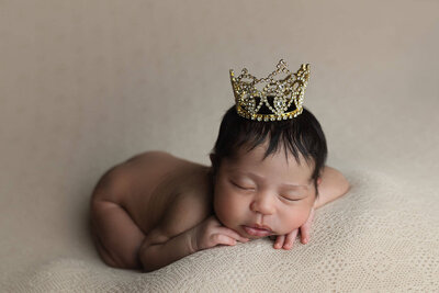 A newborn baby sleeps in froggy pose wearing only a tiny gold crown