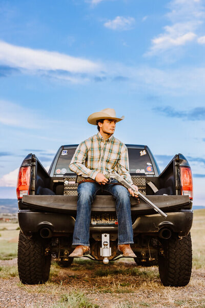 Boy in jeans and cowboy hat sits on back of truck with a gun in his hand.