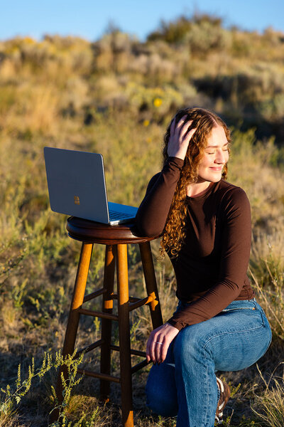 With on knee on the ground, Haylee has one hand in her hair and the other on her knee. Next to her is her laptop open on a stool.