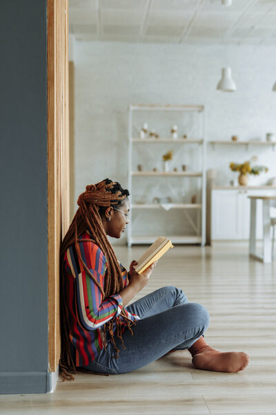 A young black woman sits cross-legged on the floor, leaning her back against the wall, reading the book in her hands. She has long dreadlocks, tied behind her head and is wearing a colorful, multi-striped button-down top, jeans, and orange socks.