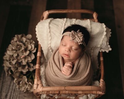 Peaceful baby sleeping on bamboo mini bed wrapped in tan with tan headband. Matching flowers to the left of  the bed.