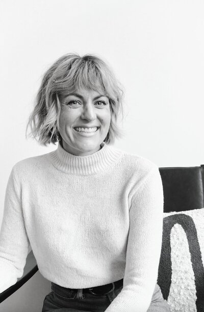 Black and white photo of 30-something woman with short, blonde hair and bangs sits and smiles at camera wearing a cozy turtleneck sweater