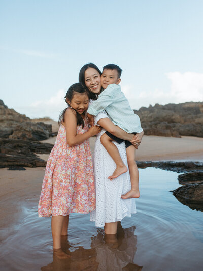 MOTHER AND CHILDREN HUGGING , THEY ARE JHOYFUL AND LOOKING AT THE CAMERA. BEACH AND WATER WITH BEAUTIFUL REFLECTIONS