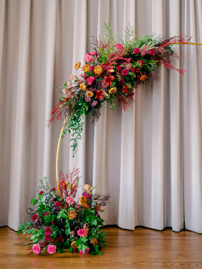 A jewel-toned floral arch sits on a stage for a wedding ceremony.