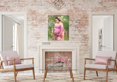 A room with a brick wall and two wooden chairs with white cushions and a glass coffee table in the center with a dusty rose floral arrangement on top and a brick filled fireplace with whitewash brick and a portrait of a pregnant women in a dusty rose lace dress standing in a field of cattails and tall grass holding her pregnant belly and wearing a pink floral headpiece in her hair hanging on the brick wall.
