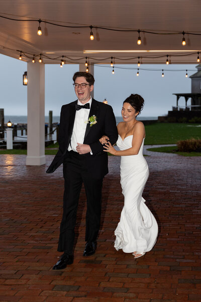New England Wedding Photographer Pricing | Kelsey Sheehan Photography Timeless Rhode Island Weddings | A couple in formal attire walks joyfully on a brick path beneath string lights. The groom laughs, and the bride smiles, exuding happiness and celebration.