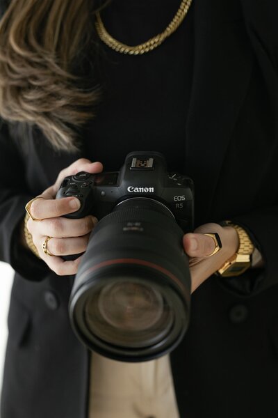 Close-up branding photo of Destination Elopement Photographer, Gaby holding a Canon EOS R5 camera, wearing a black blazer, gold jewelry, and showcasing her professional gear and personal style.