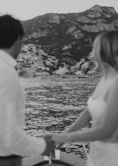 A couple holds hands while they stand on a boat overlooking Positano