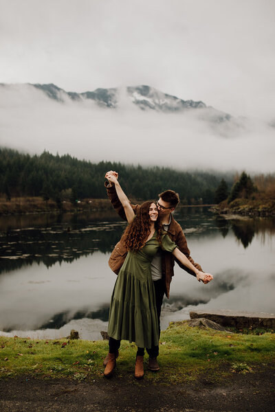man and woman stand holding hands arms outstretched with lake and mountain behind them