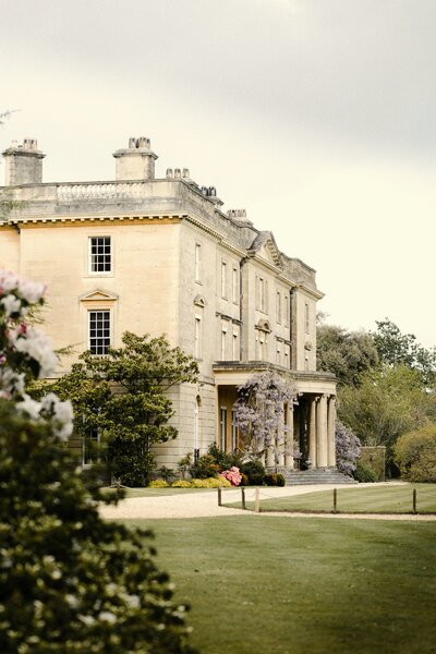 A stately country manor stands tall amidst lush green trees, its historic architecture exuding timeless elegance in the English countryside.