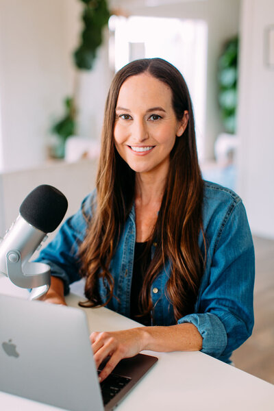 Kristie wearing a blue denim button up, sitting behind a microphone and smiling at the camera