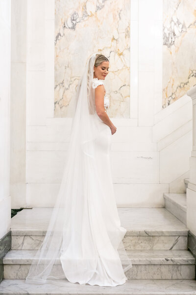 A bride in a white gown stands gracefully on marble stairs. Her long veil cascades down the steps. The backdrop features elegant marble accents, adding a classic touch to the scene.