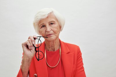 Older woman in a red blazer and gold necklace holding a pair of glasses
