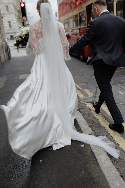 Cinematic documentary-style wedding photo by Luis Calow in Liverpool, showing a bride and groom walking together on a city street with a classic red bus in the background.
