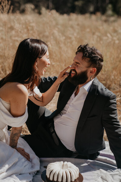 bride booping groom's nose with frosting on her finger from wedding bundt cake that is on their picnic blanket in front of them there are beige grasses and shrubs in the background