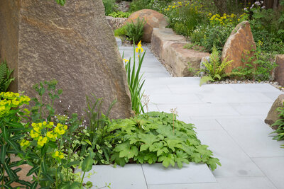 A garden pathway flanked by large stones and lush, diverse plantings, leading through a tranquil green landscape.